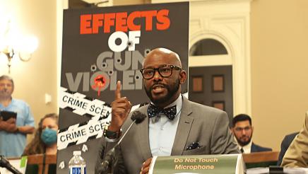 Asm. Gipson seated at microphone, speaking, with poster in background