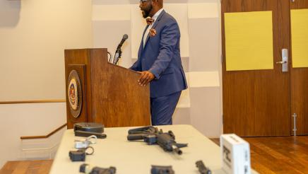 Asm. Gipson at podium, speaking beside guns on table