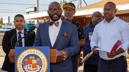 Assemblymember Gipson stands at a podium with local leaders behind him and a tavern behind them