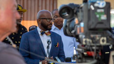 Assemblymember Gipson stands at a podium with local leaders behind him. There is a large video camera with someone controlling it in front of them.