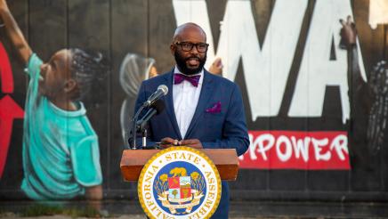 Asm. Gipson at podium, speaking, with Watts mural in background