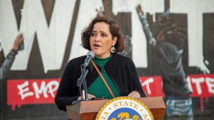 Guest speaker at podium, speaking, with Watts mural in background