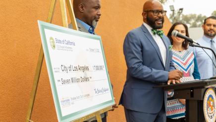 Asm. Gipson at podium, speaking, with large ceremonial check in foreground