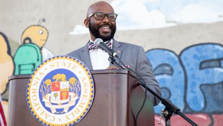 Asm. Gipson at podium, speaking, with mural in background