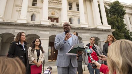 Asm. Gipson gesturing with one hand, holding papers in the other, with State Capitol in background