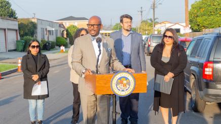 Asm. Gipson at podium, speaking, with guest speakers listening on in background