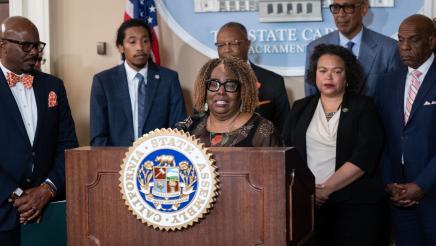 Guest speaker at podium, speaking, flanked by legislators and other guests