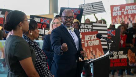 Asm. Gipson at podium, speaking, with attendees holding signs in the background