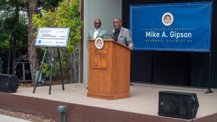 Onstage, Asm. Gipson listens on as Sen. Bradford speaks at podium 