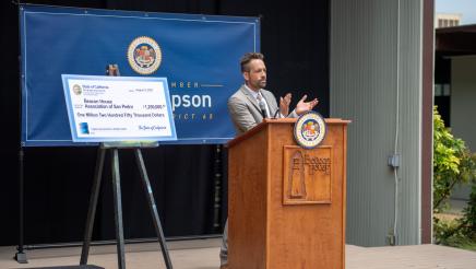  Exec. Dir. Hoggan onstage at podium, applauding, with large ceremonial check at left