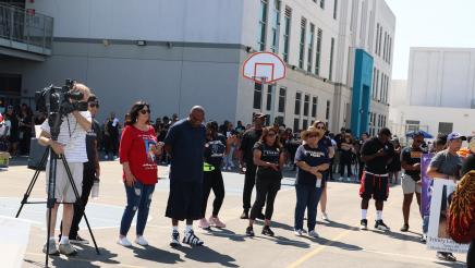 Participants staning in a line, listening to speaker, with many participants in background