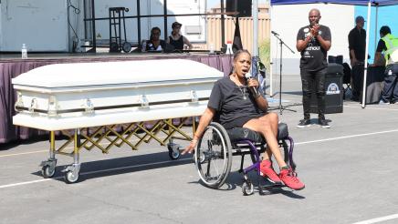 Speaker in wheelchair in front of coffin, holding microphone and speaking