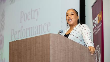 Woman at podium, speaking, with large screen in background