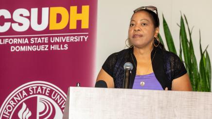 Woman at podium, speaking, with large CSUDH banner in background