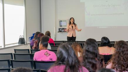 Woman standing at head of room, speaking, as audience members listen on