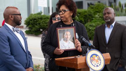 Guest speaker at podium, addressing Asm. Gipson and holding photo of victim