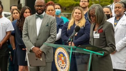 Hospital staffer at podium, speaking, with Asm. Gipson and hospital staff in background