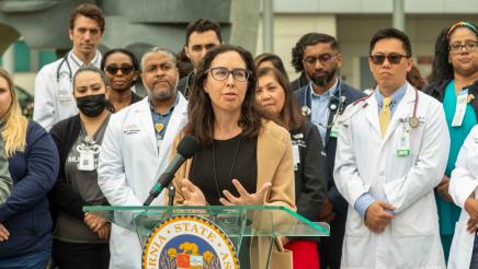Hospital staffer at podium, speaking, with other hospital staff in background