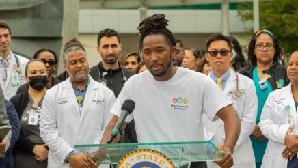 Hospital staffer at podium, speaking, with other hospital staff in background