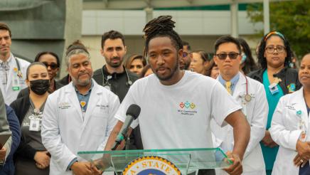 Hospital staffer at podium, speaking, with other hospital staff in background