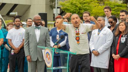 Guest speaker at podium, holding up both arms while speaking, with Asm. Gipson and hospital staff in background