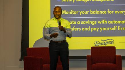 Guest speaker standing on stage, speaking, with large screen in background
