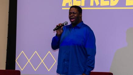 Guest speaker standing on stage, speaking, with large screen in background