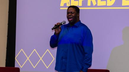 Guest speaker standing on stage, speaking, with large screen in background