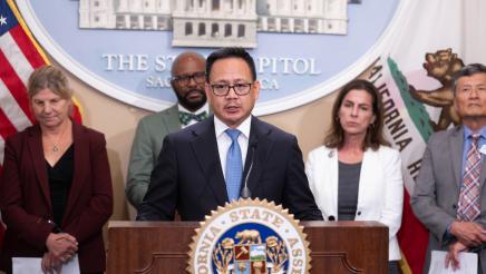 Guest speaker at podium, speaking, with large logo og the State Capitol in background
