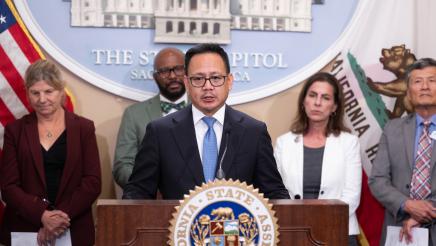 Guest speaker at podium, speaking, with large logo og the State Capitol in background