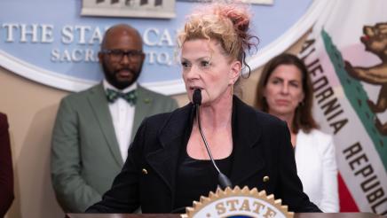 Guest speaker at podium, speaking, with large logo og the State Capitol in background