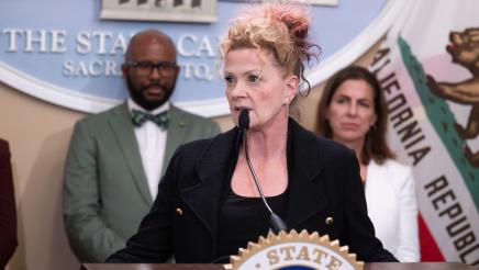 Guest speaker at podium, speaking, with large logo og the State Capitol in background