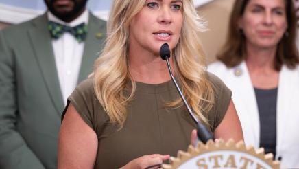 Guest speaker at podium, speaking, with large logo og the State Capitol in background