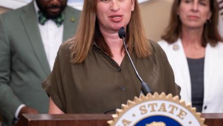 Guest speaker at podium, speaking, with large logo og the State Capitol in background