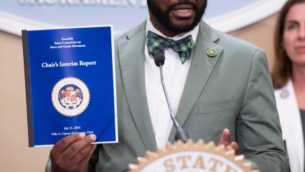 Asm. Gipson at podium, speaking and holding up report booklet