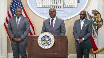 Dr. BJ Snowden at podium, speaking, as Asm. Gipson and Dr. Keith Curry listen on