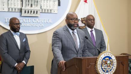 Dr. Keith Curry at podium, speaking, as Asm. Gipson and Dr. BJ Snowden listen on