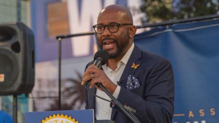 Asm. Gipson at podium, holding microphone and speaking