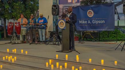 Asm. Gipson at podium, speaking, with many candles placed on steps in foreground